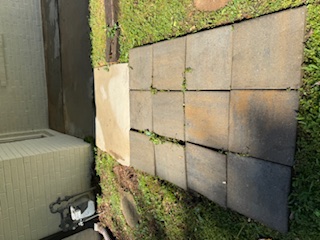 A grid of dark square concrete pavers laid on a green lawn next to a light-colored brick house wall.